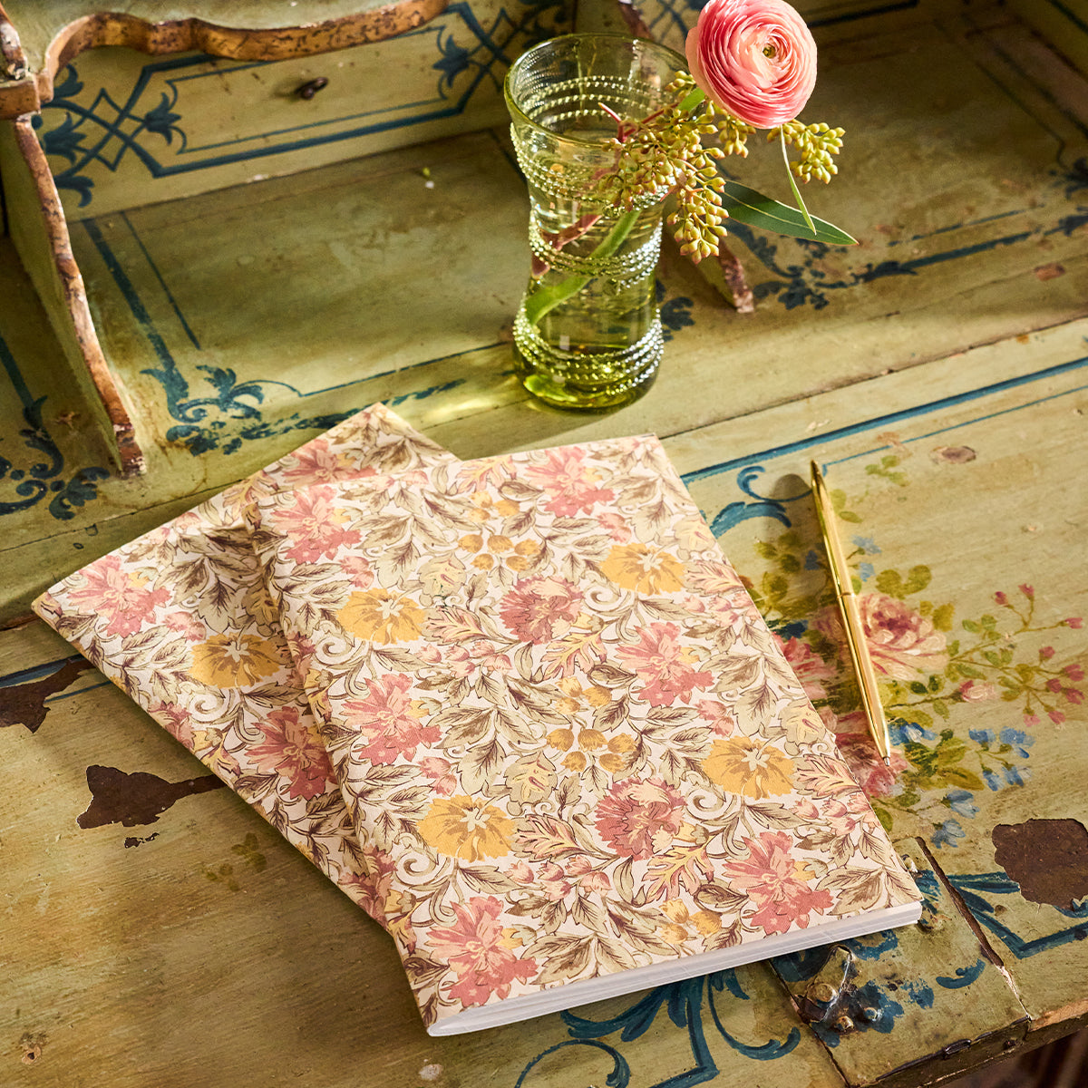 Two Willa Amber Paper Journals by Juliska, a gold pen, and a glass vase with a pink flower are arranged on a vintage, painted wooden table—a charming setting that makes these Juliska journals the perfect thoughtful gift for anyone who loves to journal.