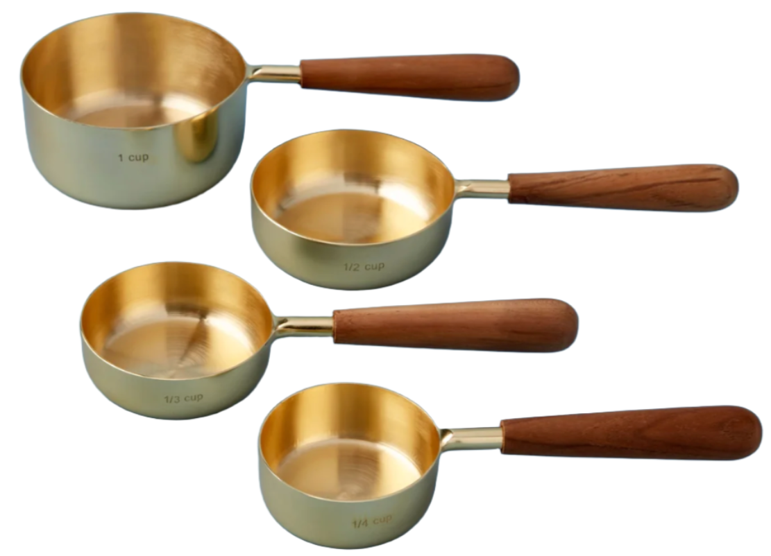 The Be Home Gold & Wood Measuring Cup Set features four handcrafted gold cups with wooden handles, labeled 1 cup, 1/2 cup, 1/3 cup, and 1/4 cup, arranged on a white background. Made in India.