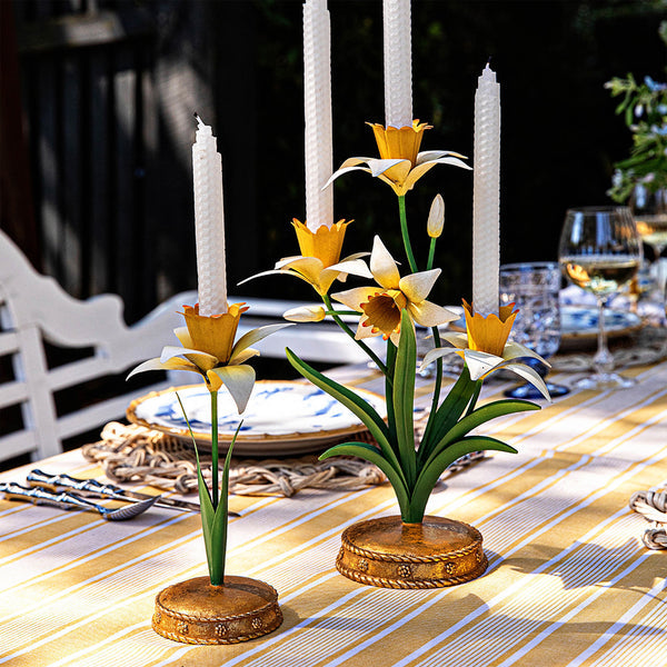 Two Juliska Meadow Walk Daffodil Candle Holder Collection pieces with tall white candles sit on a striped tablecloth, with plates, glasses, and cutlery set for a meal in the background.