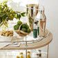 The Brays Bar Cart by Arteriors features a leather-wrapped handle and glass and wood shelves to stylishly hold a potted plant, green citrus, nuts, a silver ice bucket, cocktail shaker, and small bottle—perfect for your next cocktail party.