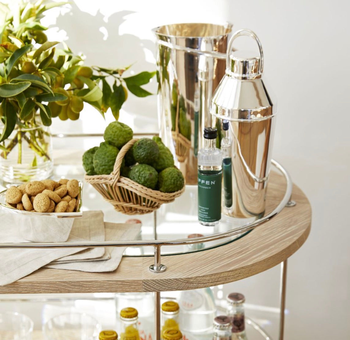 The Brays Bar Cart by Arteriors features a leather-wrapped handle and glass and wood shelves to stylishly hold a potted plant, green citrus, nuts, a silver ice bucket, cocktail shaker, and small bottle—perfect for your next cocktail party.