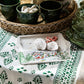 A small plate from Juliska's Berry & Thread North Pole Collection holds two powdered sugar cookies and mini candy canes, set on a green and white tablecloth beside green mugs and a wicker tray.
