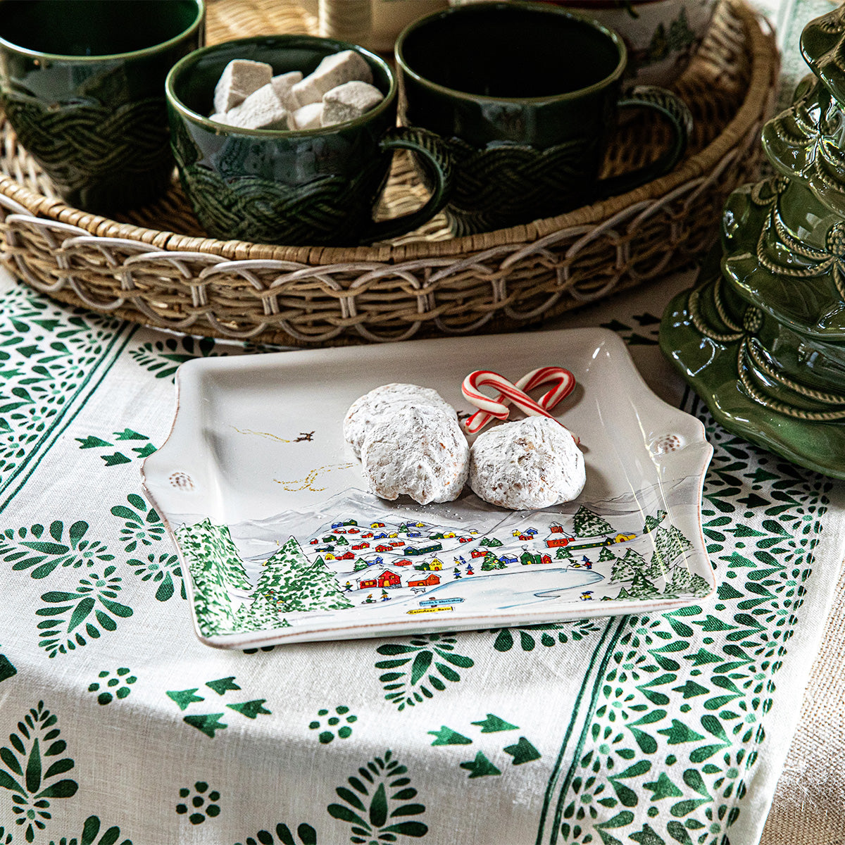 A small plate from Juliska's Berry & Thread North Pole Collection holds two powdered sugar cookies and mini candy canes, set on a green and white tablecloth beside green mugs and a wicker tray.