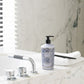 A pump bottle, black comb, and folded white towel rest on a marble bathtub edge beside modern chrome fixtures, reflecting the sophisticated Baobab Collection Gentlemen Body Wellness Collection and its amber oriental allure.