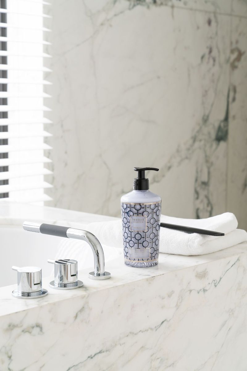 A pump bottle, black comb, and folded white towel rest on a marble bathtub edge beside modern chrome fixtures, reflecting the sophisticated Baobab Collection Gentlemen Body Wellness Collection and its amber oriental allure.