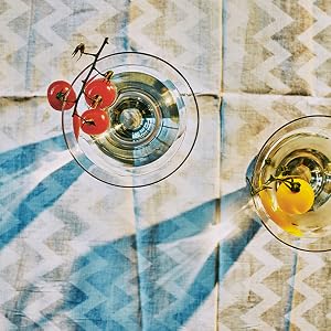 Two glasses of white wine rest elegantly on a patterned tablecloth—one accompanied by vibrant red tomatoes on the vine, the other by a golden yellow tomato—creating a scene that would surely delight Chronicle Books’ The Martini aficionados, and even cocktail expert Alice Lascelles.