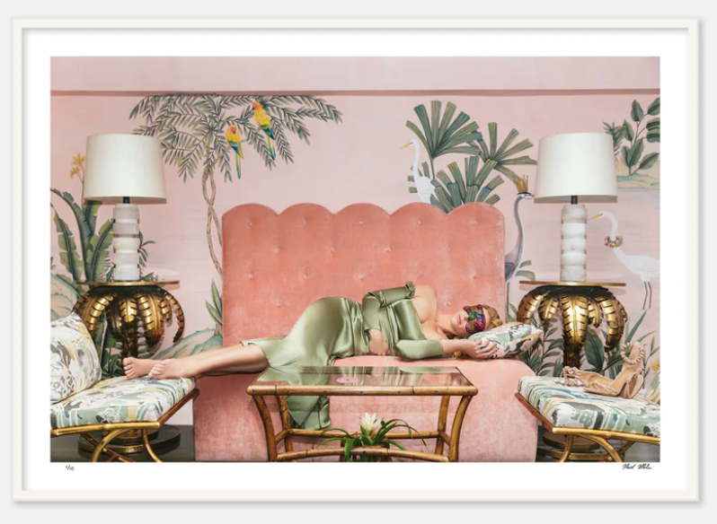 A person in a green outfit is lying on a pink sofa at the Colony Hotel, flanked by palm-shaped lamps and walls adorned with tropical plants and birds. A glass coffee table with a plant sits in front, capturing the charm of Palm Beach. This scene is depicted in Nick Mele Fine Art Nap Time by Nick Mele Photography.