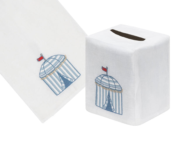 The Beach Cabana Bath Collection by Haute Home features a white Italian linen tissue box cover and towel, beautifully adorned with a hand-embroidered blue cabana design and topped with a charming red flag.