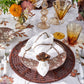 A table setting with floral-patterned plates, amber glasses, silverware, a white napkin held by the Kim Seybert Begonia Napkin Ring, and autumn-themed decor highlights natural beauty and seasonal charm.