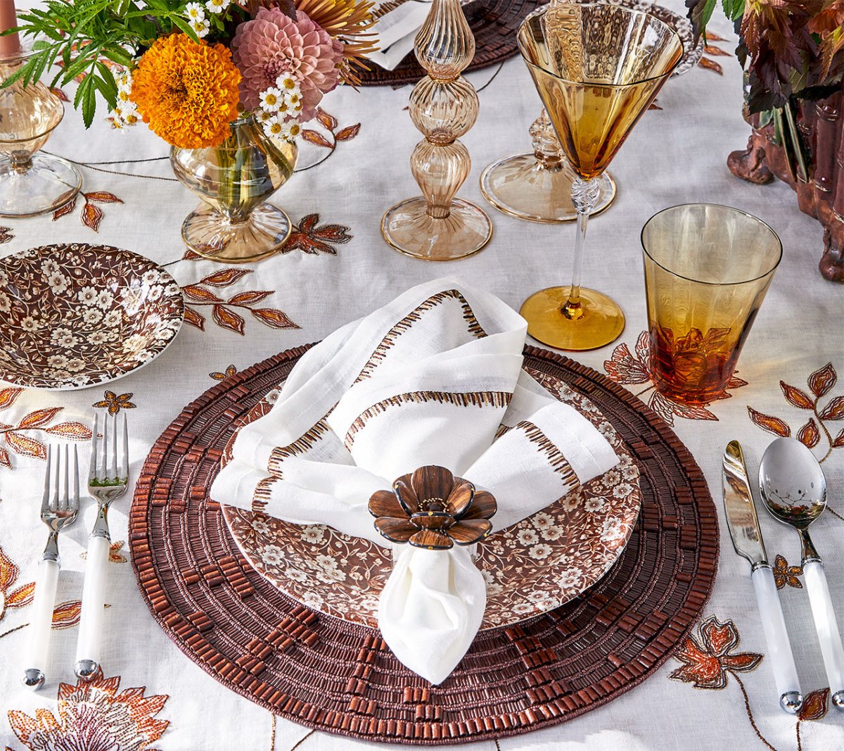 A table setting with floral-patterned plates, amber glasses, silverware, a white napkin held by the Kim Seybert Begonia Napkin Ring, and autumn-themed decor highlights natural beauty and seasonal charm.