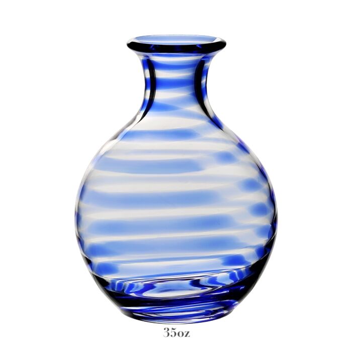The Bella Blue Carafe Collection by William Yeoward Crystal features a round glass carafe with blue and clear horizontal stripes, a narrow neck, and flat base. Holds 35 oz., ideal for wine. Proposition 65 warning: may contain harmful materials.
