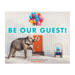 An elephant carries colorful balloons in its trunk toward an orange door at the Parker Palm Springs, passing a luggage cart and the Be Our Guest! sign on a white patterned wall.