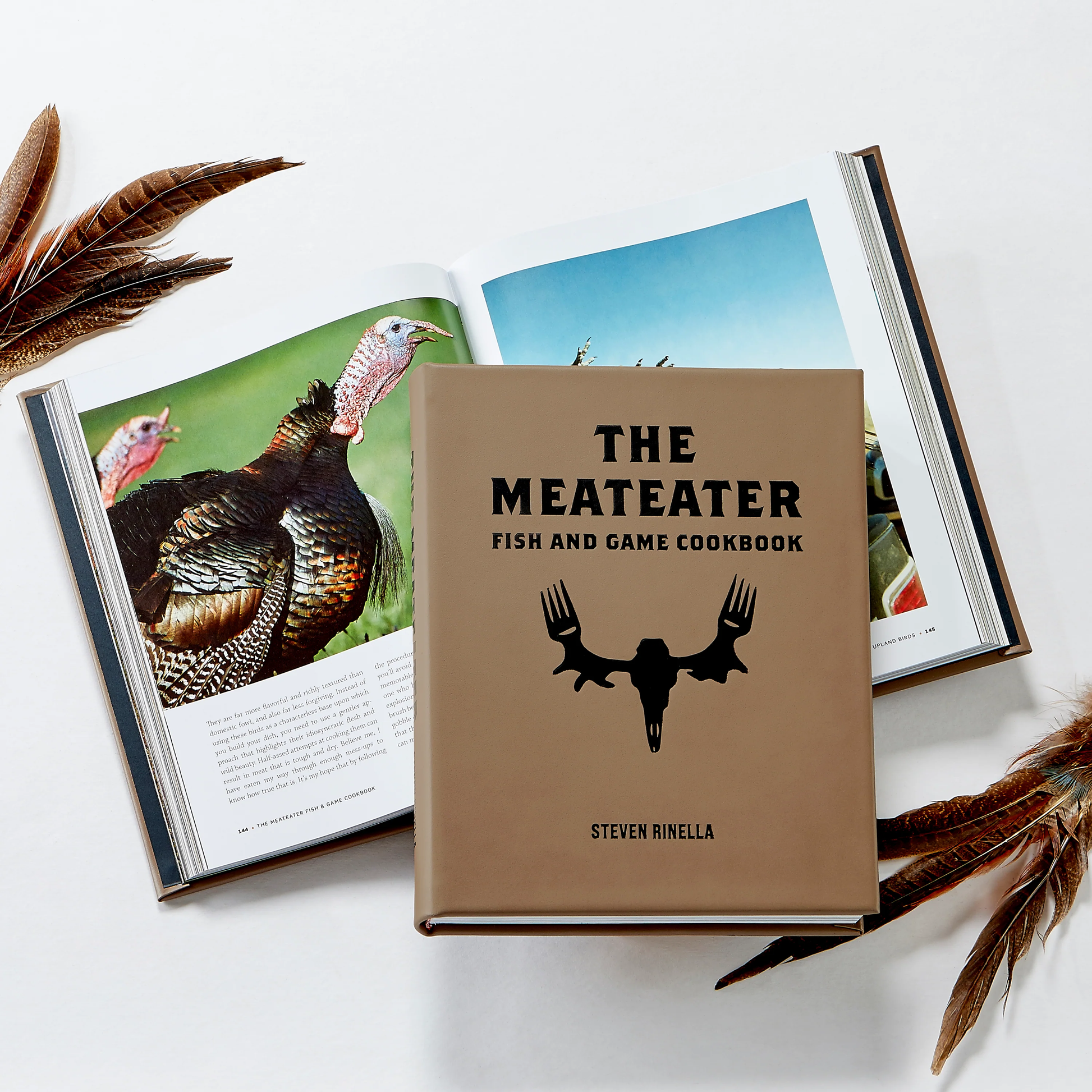 The Meateater Fish and Game Cookbook by Graphic Image is placed atop an open book with turkey images, accented by nearby feather decorations.