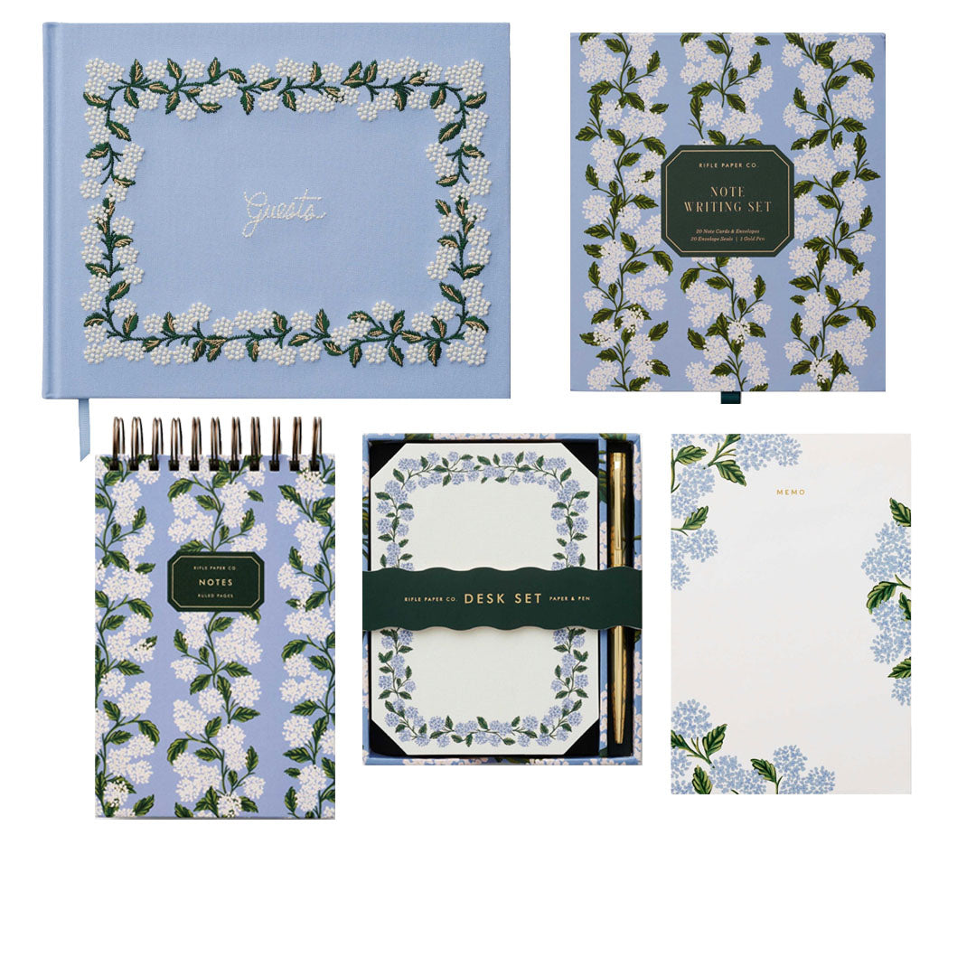 The Blue Hydrangea Desk Accessory Collection by Riffle Paper Co. features a hydrangea notepad, letter writing set with envelopes and pencil, journal, and memo pad—all adorned with elegant white and green floral designs.