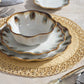 From Blue Pheasant’s Amoria Dinnerware Collection, a white ceramic bowl and two brown-edged plates with vintage charm are stacked on a round woven placemat atop a wooden table with cutlery beside them.