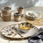 A ceramic platter of oysters on ice, Amoria Dinnerware Collection by Blue Pheasant, lemon wedges, woven glasses, stacked plates with forks, and a blue-striped napkin on beige evoke vintage-inspired elegance.
