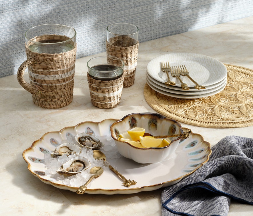 A ceramic platter of oysters on ice, Amoria Dinnerware Collection by Blue Pheasant, lemon wedges, woven glasses, stacked plates with forks, and a blue-striped napkin on beige evoke vintage-inspired elegance.