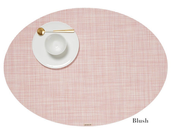 The Chilewich Oval Placemat Collection features a pink placemat with a Mini Basketweave texture, beautifully arranged with a white plate, bowl, and gold-handled spoon to add subtle sophistication to any dining occasion.