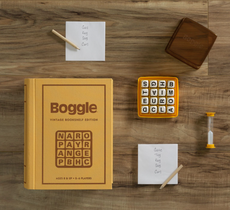The Boggle Vintage Bookshelf Edition by WS Game Company is displayed with letter cubes, two pads with written words, a sand timer, and a wooden lid. The book-style box reads "Boggle: Vintage Bookshelf Edition," paying homage to the vintage charm of Parker Brothers' 1976 edition.
