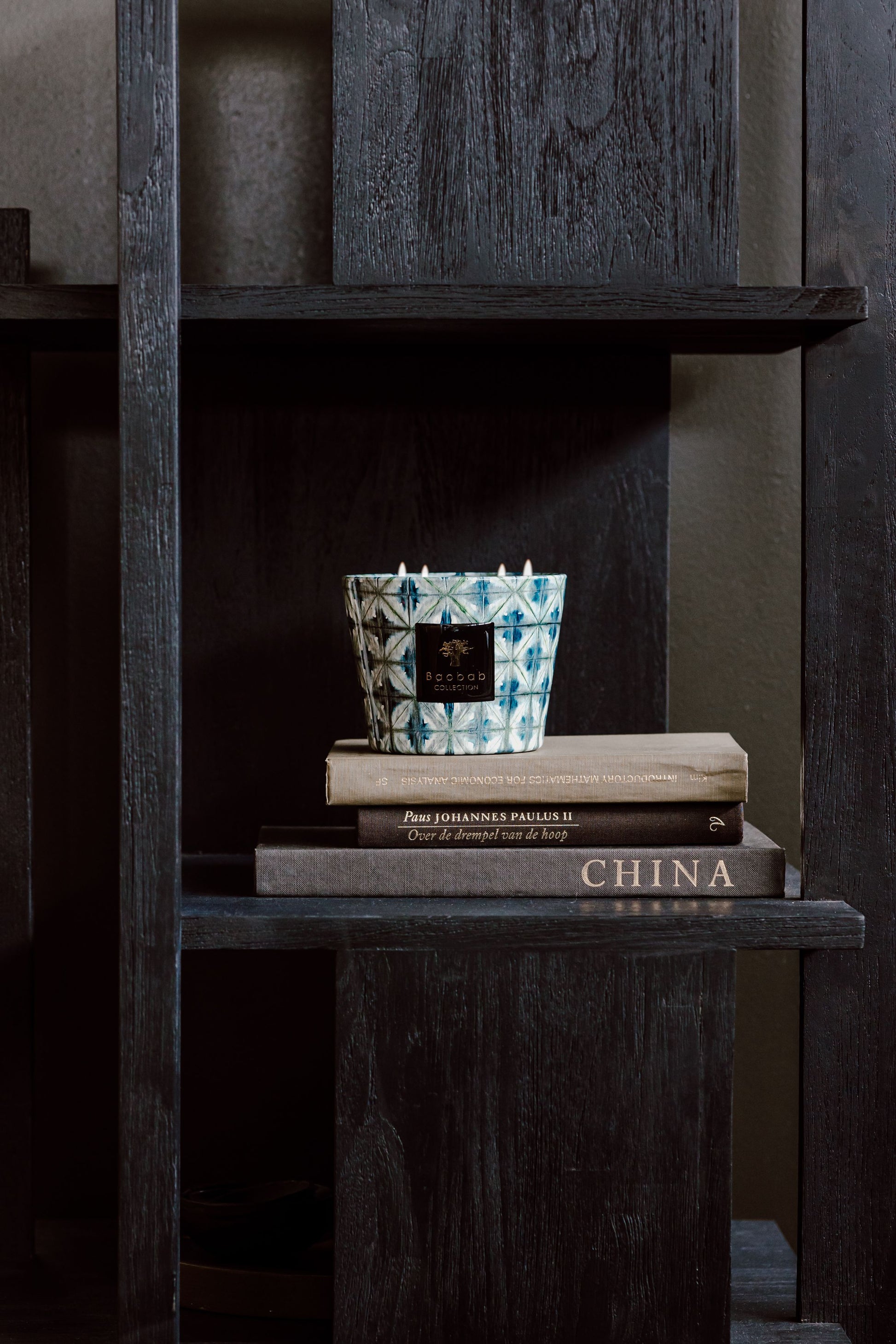 A ceramic mug and two stacked books titled "Johannes Dahlem" and "China," next to a Baobab Collection Bohomania Kilan candle on a shelf in a dark wooden bookcase.