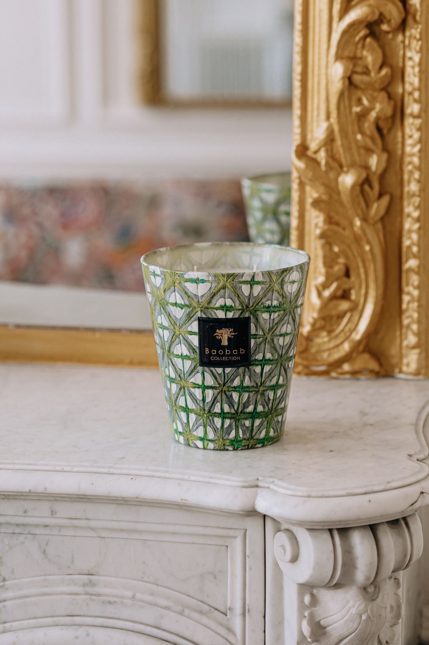 A Baobab Collection Bohomania Lazlo scented candle in a cut glass holder placed on a white marble mantel, with a gilded mirror and ornate decor in the background.