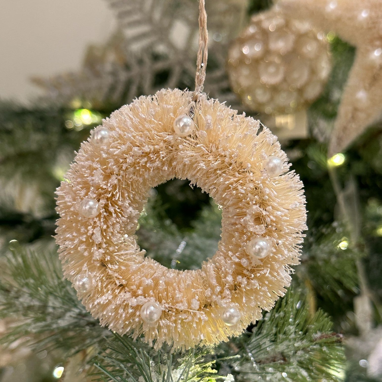 A close-up of a Dekorasyon Mini Bottlebrush Wreath Ornament, cream-colored with white beads, hanging on green branches with blurred holiday decorations in the background.