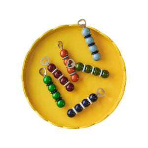 Six vibrant bead caterpillar bottle openers from the Addison Ross Bobbin Bottle Opener Collection, each with a bottle cap head, are arranged on a round yellow tray against a white background—creating a stylish gift or quirky decorative accent.