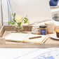 A wicker tray holds a notebook, pen, Juliska Clever Creatures Whitewash Honey Pot with a wooden spoon, a jar of clotted cream, mug, and small vase with white rose on a bed dressed in white and blue linens.