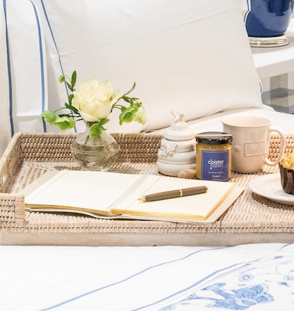 A wicker tray holds a notebook, pen, Juliska Clever Creatures Whitewash Honey Pot with a wooden spoon, a jar of clotted cream, mug, and small vase with white rose on a bed dressed in white and blue linens.