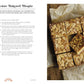 A tray of white chocolate bakewell blondies—taking inspiration from delicious recipes featured in *The Brownie Book by Kate Jenkins* from Penguin Random House—is cut into squares and topped with flaked almonds. The blondies rest on parchment paper atop a rustic wooden surface.