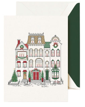 The Crane Cozy Brownstones Card Set features festive townhouses decorated with wreaths and garlands, flanked by Christmas trees, with a green envelope partially visible behind this charming holiday card.