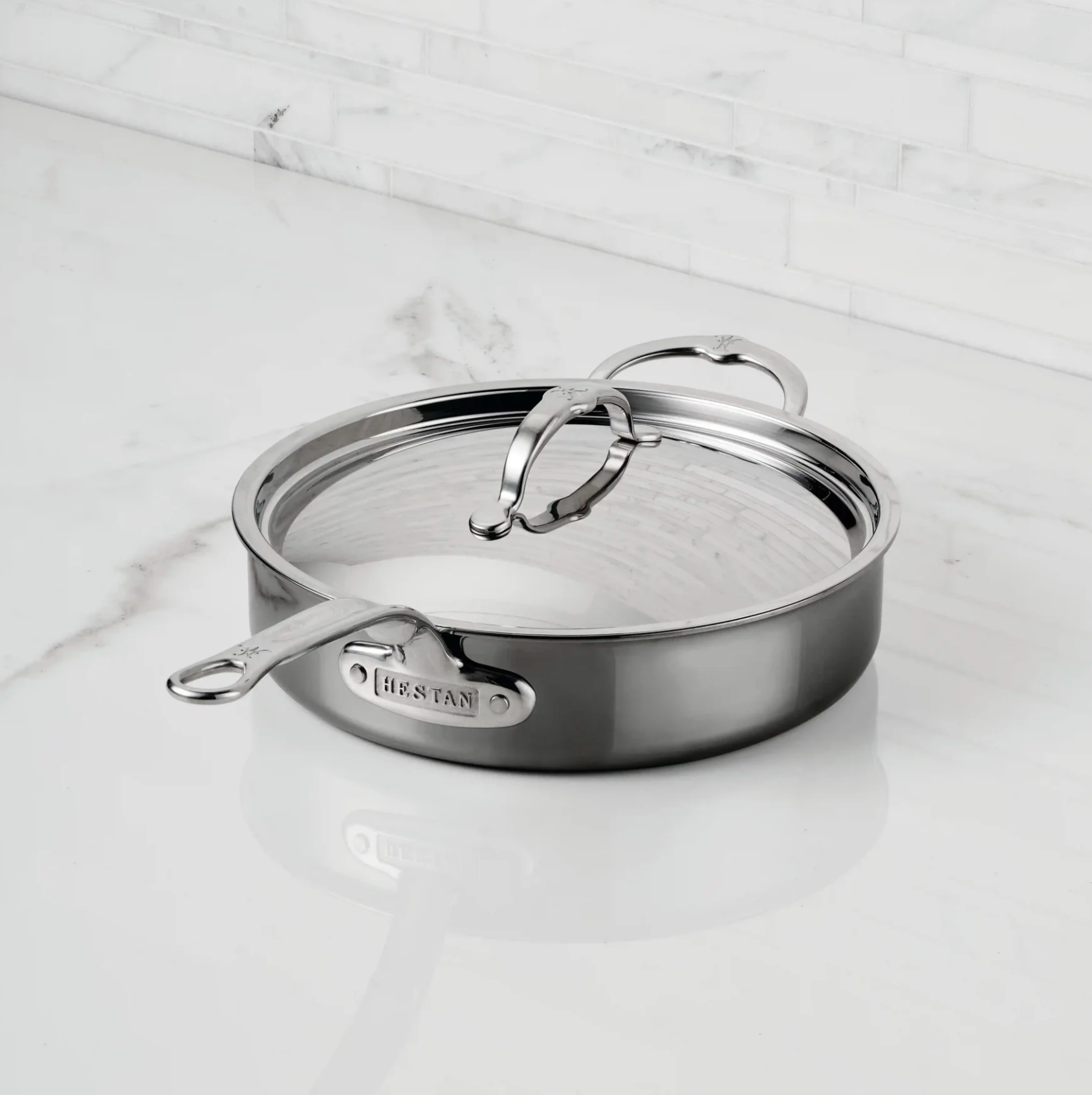 A Hestan NanoBond Covered Sauté pan sits on a white marble countertop against a white tiled backsplash.