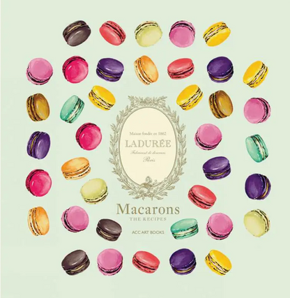 An assortment of vibrant macarons is elegantly arranged in a circle around the cover of Simon & Schuster’s Ladurée Macarons, which features a decorative central emblem, showcasing the artistry behind crafting exquisite ganache-filled delights.
