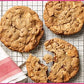 Three large cookies, inspired by Martha Stewart’s Cookie Perfection from Penguin Random House—one broken in half—rest on a wire cooling rack beside a red and white striped napkin.