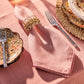 Draped in sunlight, the Berry Scallop Napkin from Juliska, accented with a decorative ring and placed between a fork and vintage floral plates, rests elegantly atop matching Berry Scallop woven linen tablecloths—an inviting scene to showcase your favorite pieces from the Juliska collection.