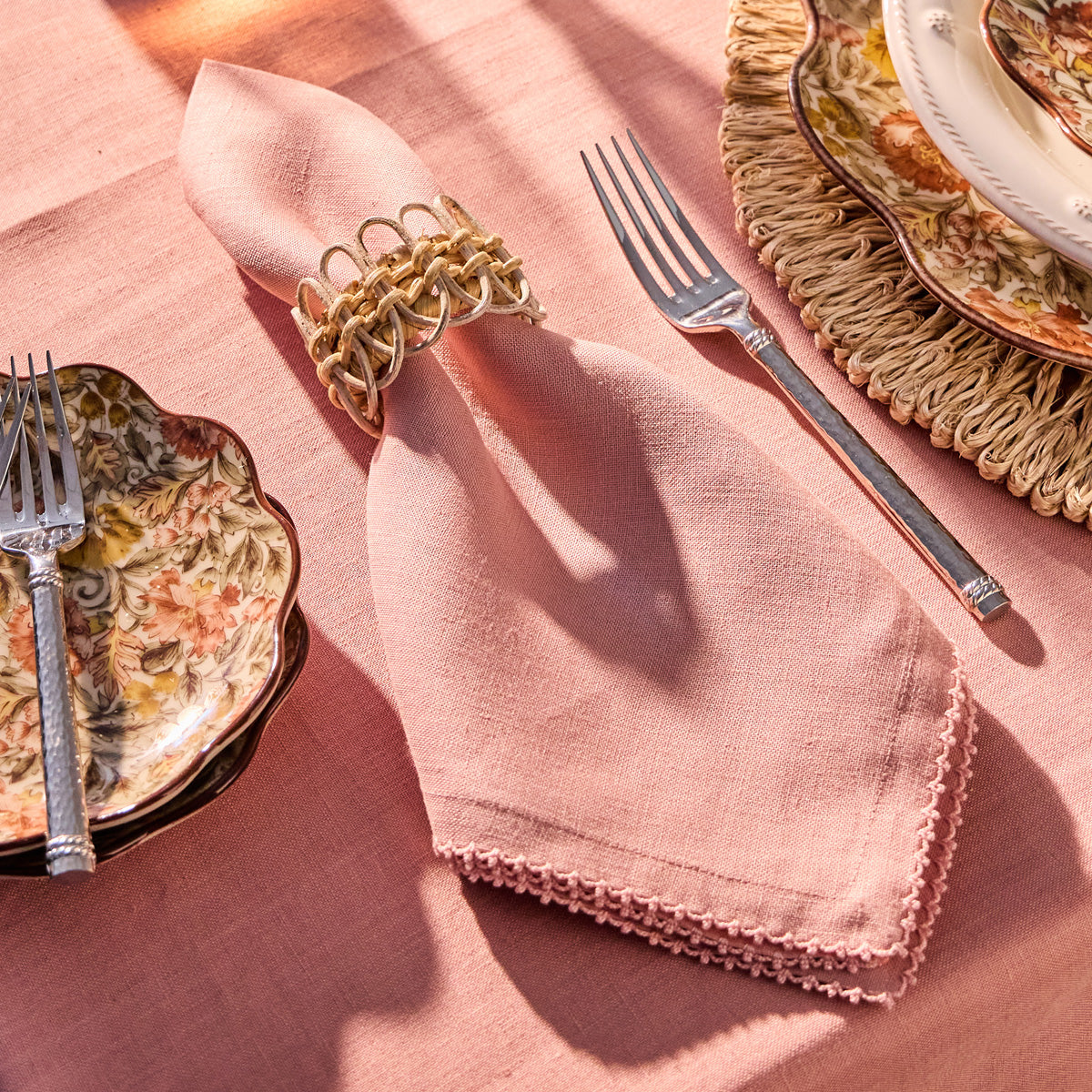 Draped in sunlight, the Berry Scallop Napkin from Juliska, accented with a decorative ring and placed between a fork and vintage floral plates, rests elegantly atop matching Berry Scallop woven linen tablecloths—an inviting scene to showcase your favorite pieces from the Juliska collection.