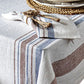 The Danish Stripe Table Linen Collection by Juliska, inspired by Scandinavian design, features a linen tablecloth with blue and brown stripes, draped over a corner and paired with a matching napkin and woven placemat.