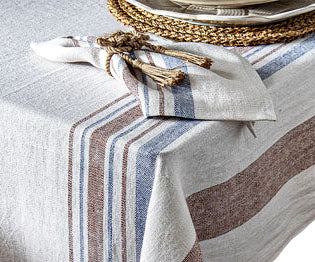 The Danish Stripe Table Linen Collection by Juliska, inspired by Scandinavian design, features a linen tablecloth with blue and brown stripes, draped over a corner and paired with a matching napkin and woven placemat.