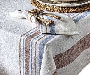 The Danish Stripe Table Linen Collection by Juliska, inspired by Scandinavian design, features a linen tablecloth with blue and brown stripes, draped over a corner and paired with a matching napkin and woven placemat.