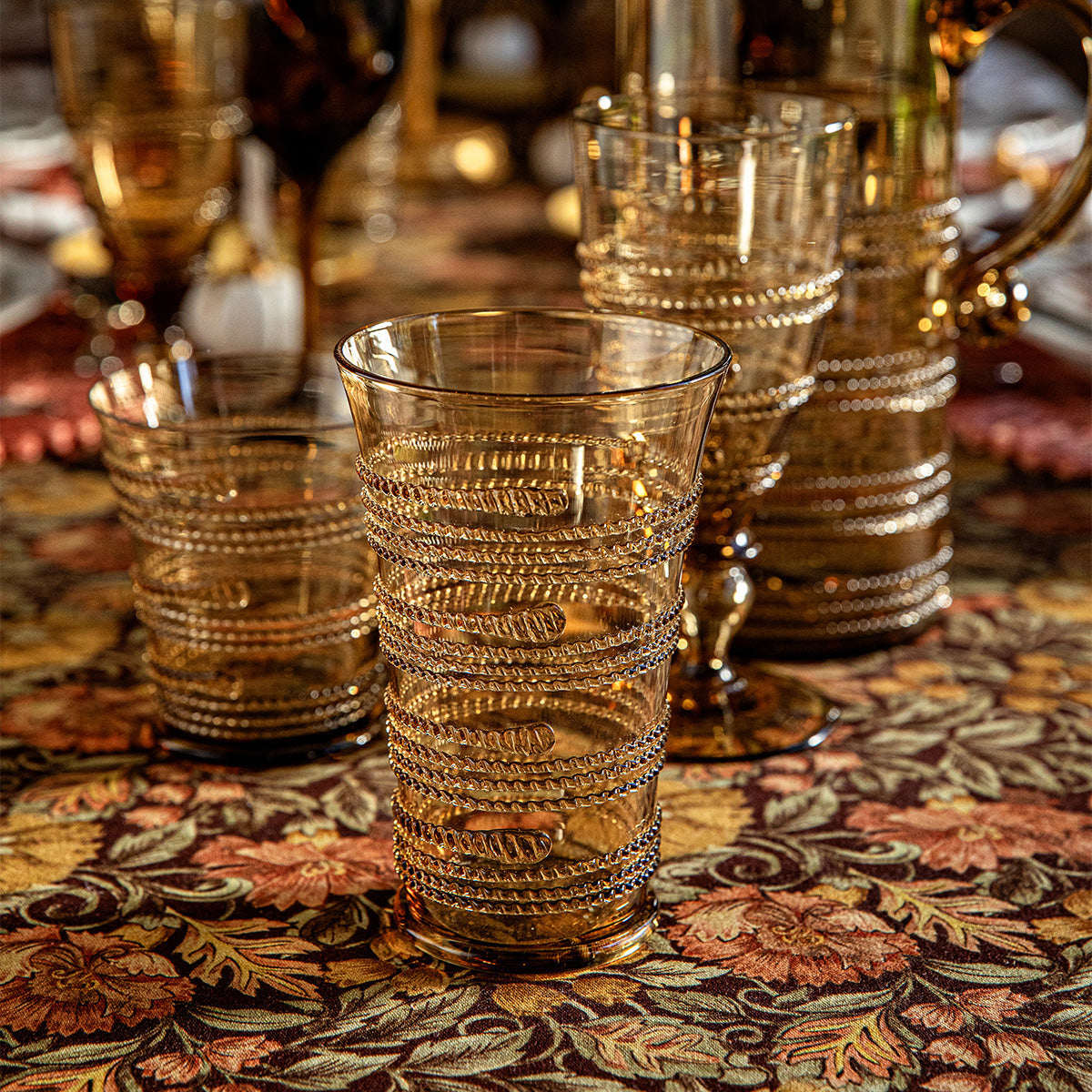 Four gold-tinted glasses from the Ella Drinkware Collection by Juliska, featuring artisan spiral bead patterns, are arranged on a floral-patterned tablecloth, with additional Juliska glassware and table settings visible in the background.