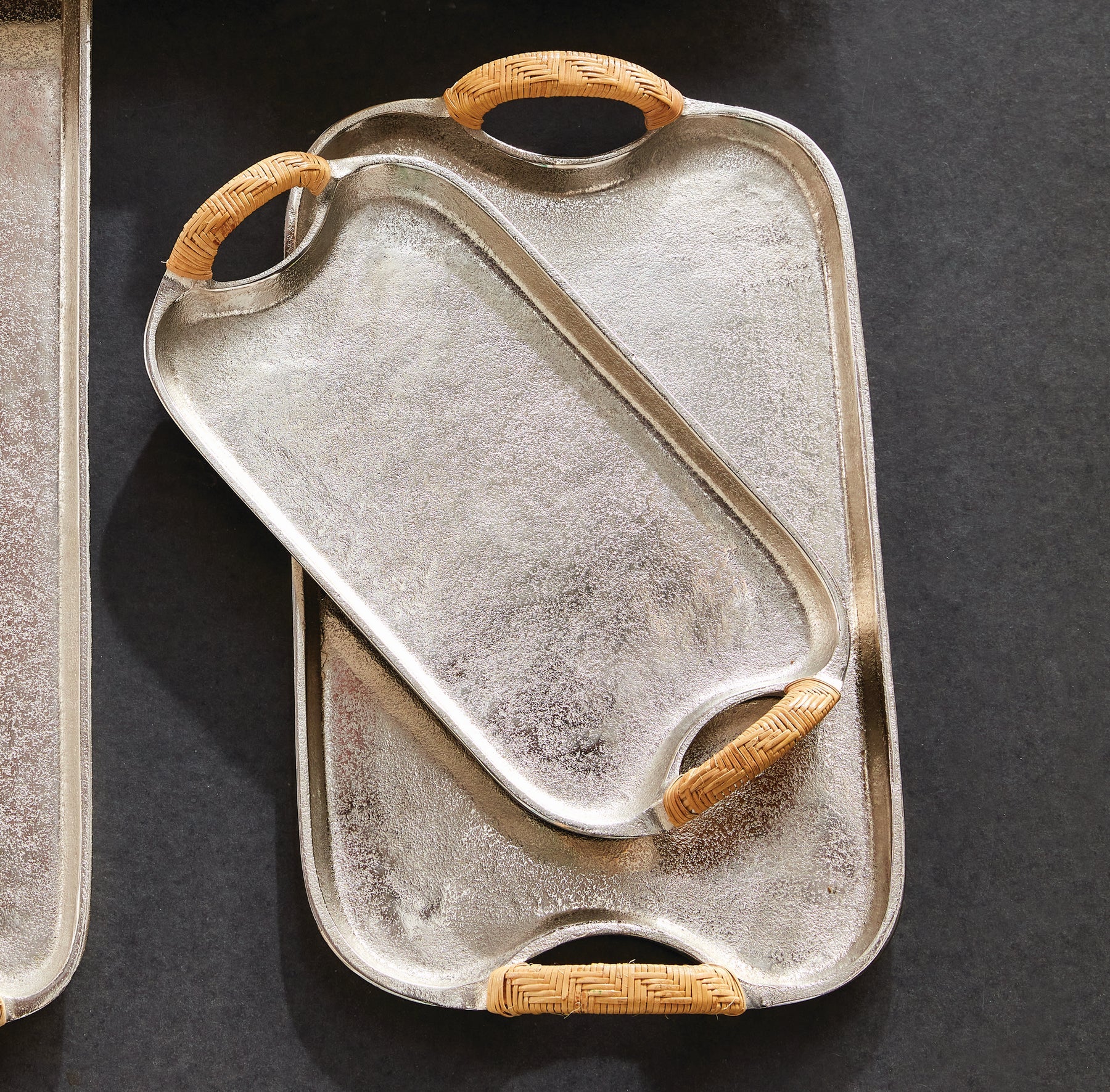 Two modern trays from the Helena Decorative Tray Collection by Napa Home & Garden, crafted in cast aluminum with elegant rattan-wrapped handles, are stacked on a dark surface with the smaller nested inside the larger.