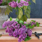 A bunch of purple lilac flowers with green leaves lies on a wooden table next to pruning shears and a vase filled with more lilacs, embodying the monochromatic bouquet style featured in *Color Me Floral* by Chronicle Books.