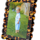 A young child in a light blue outfit stands on grass outdoors, pictured in Tara Wilson Designs' Scallop Frame Collection, featuring a scalloped, tortoiseshell-patterned border.