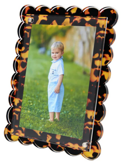 A young child in a light blue outfit stands on grass outdoors, pictured in Tara Wilson Designs' Scallop Frame Collection, featuring a scalloped, tortoiseshell-patterned border.