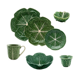 The Green Cabbage Dinnerware Collection by Bordallo Pinheiro features plates, bowls, a mug, and small dishes, crafted in Portugal and shaped like cabbage leaves.