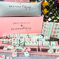 A Bespoke x Gray Malin Mahjong Collection set with tiles featuring colorful dogs, travel icons, and landmarks, displayed in front of a pink storage box and a light blue carrying pouch.