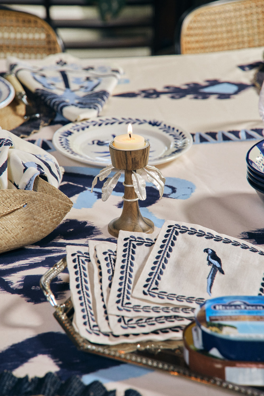A lit candle on a wooden holder sits beside patterned plates, a can of fish, and the Johanna Ortiz Caica Cocktail Napkin Set, creating an atmosphere of vibrant elegance.