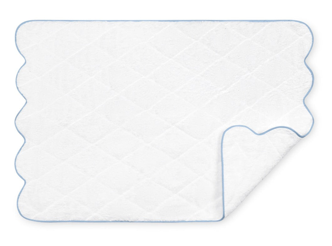 The Matouk Cairo Scallop Bath Collection's white quilted bath mat flaunts a scalloped edge with blue trim and a partially folded corner to reveal its luxurious texture. Crafted from soft, long-staple cotton, it perfectly complements our terry towels.