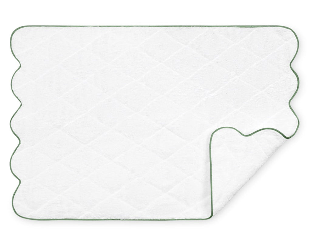 The Matouk Cairo Scallop Bath Collection mat features a luxurious white quilted design with a scalloped edge and green trim, crafted from long-staple cotton, partially folded at one corner.
