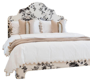 The EJ Schrader Pyne Hollyhock King Bed features a floral-patterned upholstered headboard and base, white bedding, beige and white pillows, and a beige throw blanket.